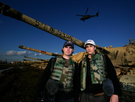 Two men in camouflage vests pose in front of a military tank.