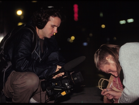 A man with headphones and holding a camera films another person from up close.