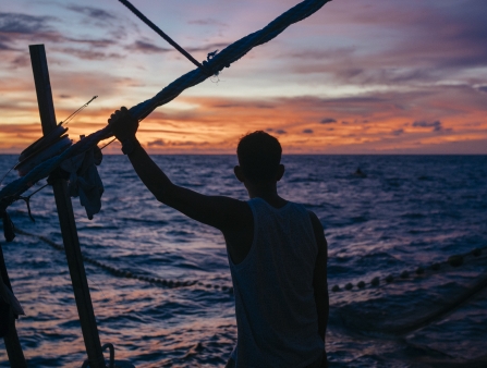 A man leans against a pole while the sun sets on the sea.