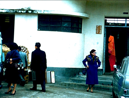 A woman in a blue outer coat stands in front of a white-walled house.