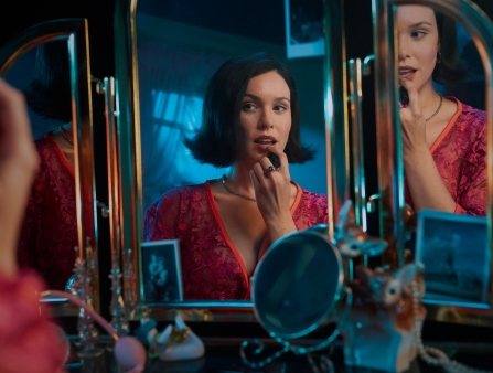 A reflection in a vanity mirror of a woman with a short brown bob putting on lipstick.