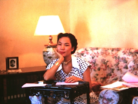 A young woman with pinned back black hair and a fashionable capped sleeve dress sits on a floral sofa, leaning on a small writer's desk.