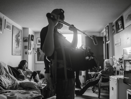 A sound recorder holds a boom mic to record a conversation between a woman and an older man in a living room.