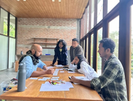In a bright room with floor to ceiling windows, five individuals are gathered on a table looking over a number of papers