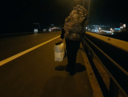 At night, a blurry image of a person walking along the side of a highway, wearing a large camo backpack.