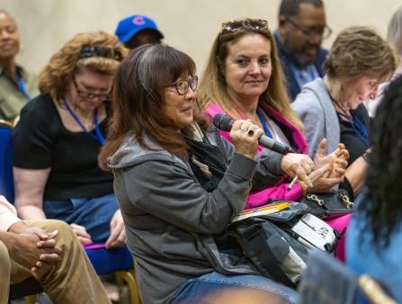 A woman with glasses and long dark hair holds a mic surrounded by fellow seated audience members