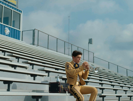 On school bleachers, a mariachi player dressed in yellow plays his trumpet.