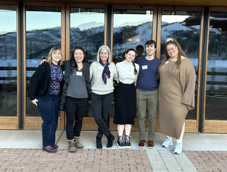 Six smiling people stand outside. Snowy mountains are reflected in the glass doors behind them.
