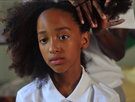 A young Black girl sits while her hair is gathered into a pigtail.