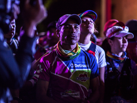 A brown-skinned man wearing athletic gear and a cap is surrounded by other runners in a nighttime shot.