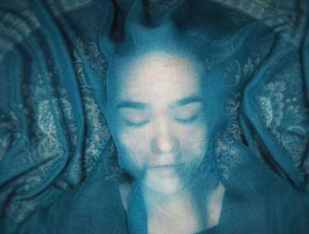 Under a blue cloth that is semi-transparent, a teenaged girl lays on the floor with her eyes closed.