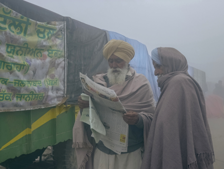 Two men wearing yellow turbans and gray shawls read a newspaper.