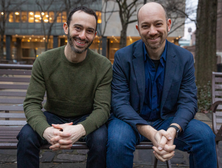 Two bearded men (one in a green sweater and jeans; the other in blue blazer, blue shirt and jeans) sit on a bench and smile at the camera