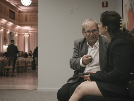 An older man in a blazer and white shirt sits on a bench next to a younger woman in a black skirt and blazer, her dark long hair pulled back with a clip