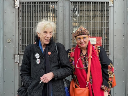 An older white woman with white hair in a black winter jacket stands next to a woman in a bright-colored ensemble in front of the Anthology Film Archives building