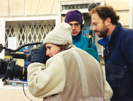 A white woman in a winter coat and hat, stares into the lens of a camera while two men (one in a purple beanie, another in a blue jacket) stand nearby.
