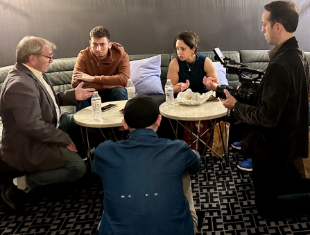 Three men and a woman sit around two small tables as a cameraman captures their conversation