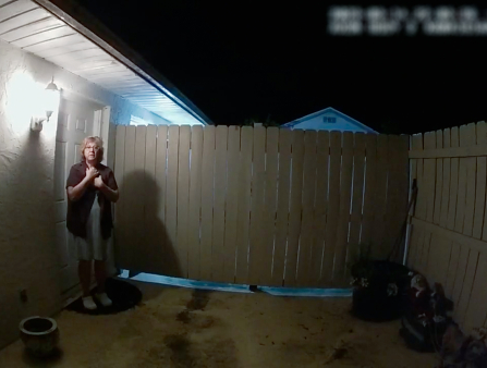 Bodycam still of a white woman with glasses and light shoulder-length hair standing in front of her front door at night.
