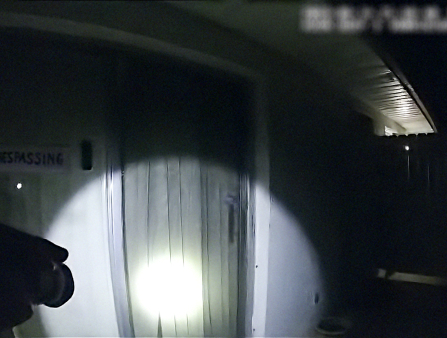 Dark, bodycam still of a glass door at night lit by a flashlight hoisted to a gun.