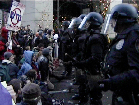 Grainy shot of police in riot gear facing off seated, peaceful people protesting the WTO