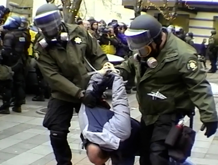 Two police officers with protective helmets on drag a zip-tied protestor away