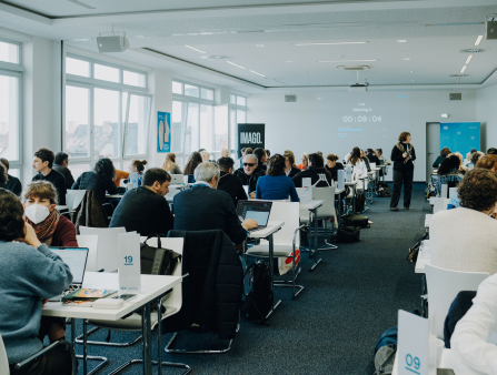 An office-like space with bright windows is filled with people sitting down for meetings.