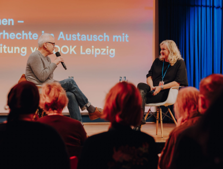 A white man with white hair and black-rimmed glasses in jeans and a sweater sits at a stage with a mic next to a white woman with white blond hair, all in black.