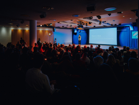 A woman on a stage holding a microphone leads a talk to an auditorium full of people