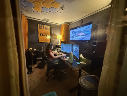 A white woman in a t-shirt sits at an editing bay surrounded by screens