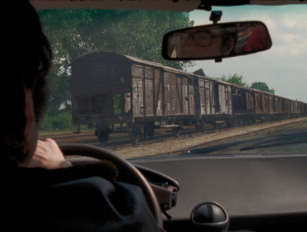 Shot from inside a car, we see a man in profile driving right by a train