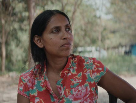 A brown woman wearing a bright, floral short-sleeved shirt, with long dark hair tied behind her back stares into the distance.