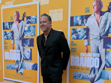 A white man with grey hair wearing a black suit and black shirt poses in front of a step and repeat