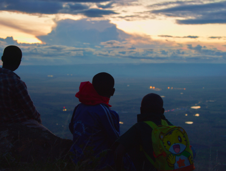 Three young Black boys seen from behind, one wearing a cartoon-decorated yellow backback, look out in to the fields and the skies in front of them