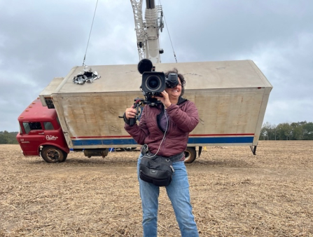 A white woman in jeans and a jacket holds a camera out on a field as a truck behind her is rigged for a stunt.