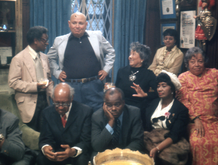 Grainy 1970s photo of a group of older Black men and women posing for a photo in someone's living room