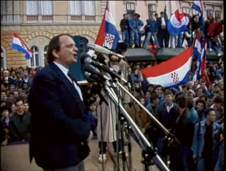 A white man in a suit in front of a handful of microphones, addresses an outdoor crowd that flies various Croatian flags all around them