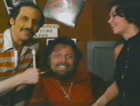 Grainy image of a white man with short dark hair and a mustache, wearing a striped polo shirt holding a mic next to an older tan man in a red shirt and a young white woman with short black hair and a sleeveless black shirt