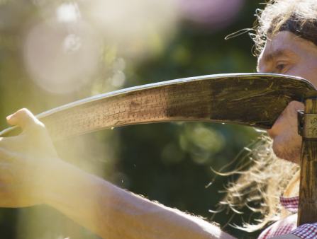 A man with long hair tied behind his back holds a scythe with care