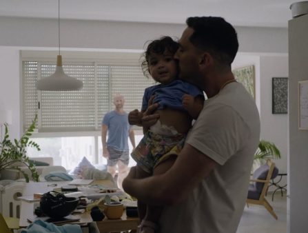 A young brown-skinned man hugs and kisses a young toddler girl amid a slightly messy living room while a balding white man in shirt and shorts looks at them in the background