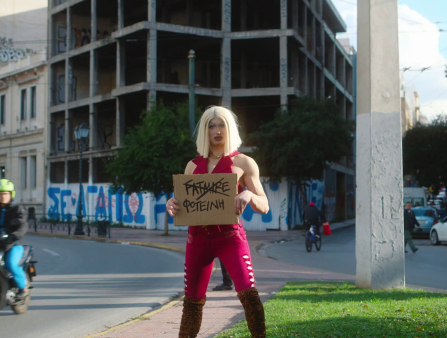 A slim white drag queen with a blond bob and tight red outfit stands out on the streets holding a sign