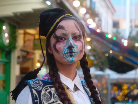 A young woman with outrageous blue, white, and pink face paint and braids stands outside in the street