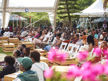 An outdoor row of seats is filled by Black folks of all ages, many of them wearing colorful shorts; a pink flower can be seen blurry in the foreground