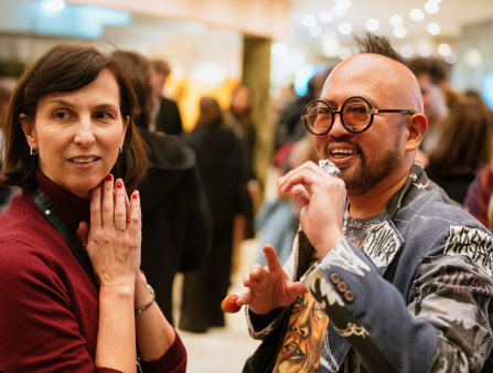 A white woman with shoulder-length dark hair in a maroon cardigan stands next to a balding man with dark-rimmed glasses wearing a funky jacket.