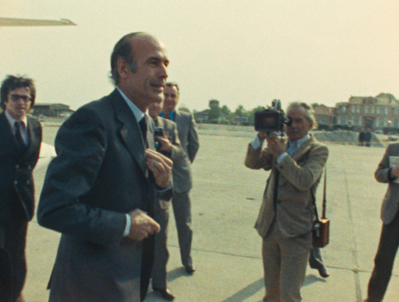 A balding white man in a suit greets photographers outside of an airplane