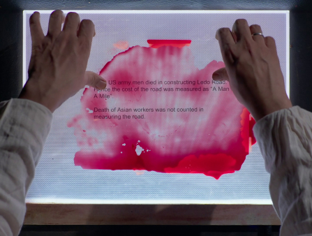 Two hands can be seen holding up a document that reads "US army men died in constructing Leto Road" against a red-splattered overlay with light coming up from below