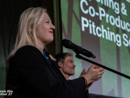 A blond white woman with shoulder-length hair wearing a black blazer claps on stage next to a microphone with film screen behind her