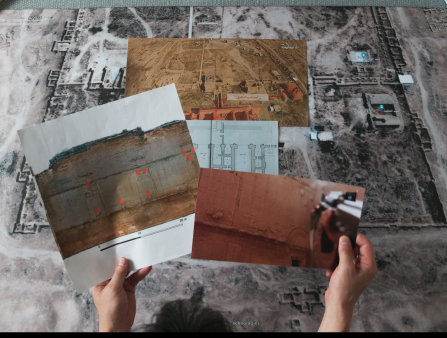 Shot from above, a man's hands hold two photos of walls and buildings, while a bird's eye view of a property is seen in the floor in the background