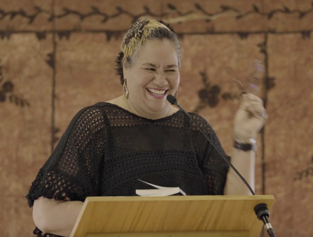 A heavy-set brown-skinned woman with short dark hair that has blond streaks stands at a lectern with a mic, wearing a black shirt