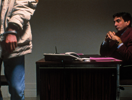 A man in handcuffs is seen in foreground from below the shoulders; another man sitting at a desk