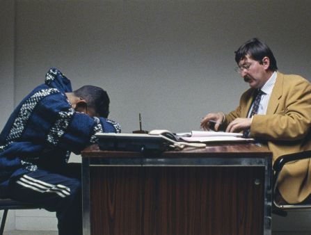 A man is hunched over a desk while a man with a mustache in a mustard blazer looks at him